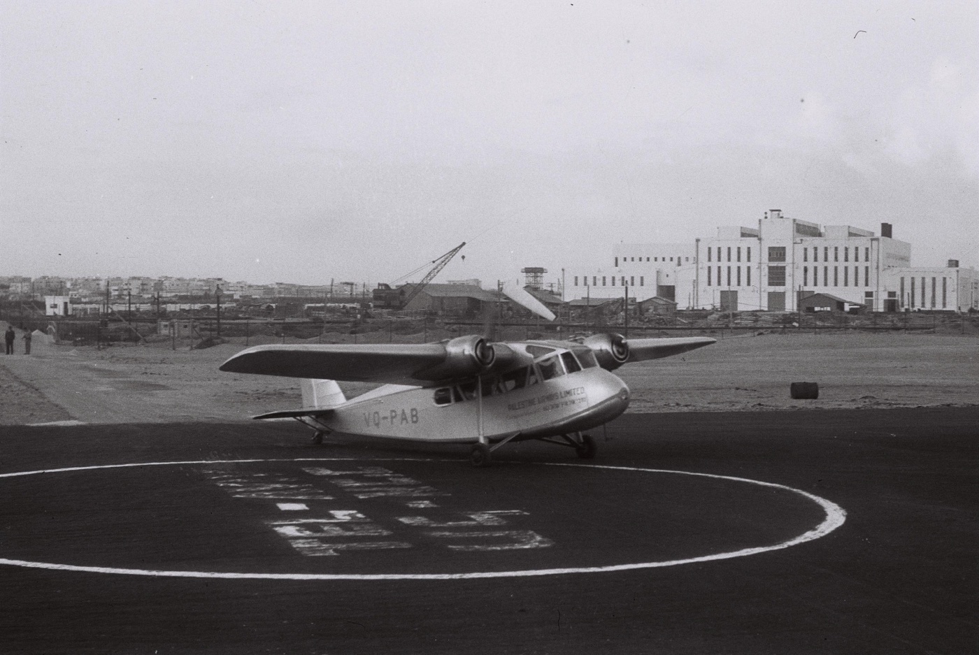 Short Scion VQ-PAB of Palestine Airways, PALESTINE AIRWAYS LIMITED lettered on the fuselage, taxiing on the circle of the Tel Aviv airfield in 1938; behind the field stands the new white Reading power station of the Palestine Electric Company