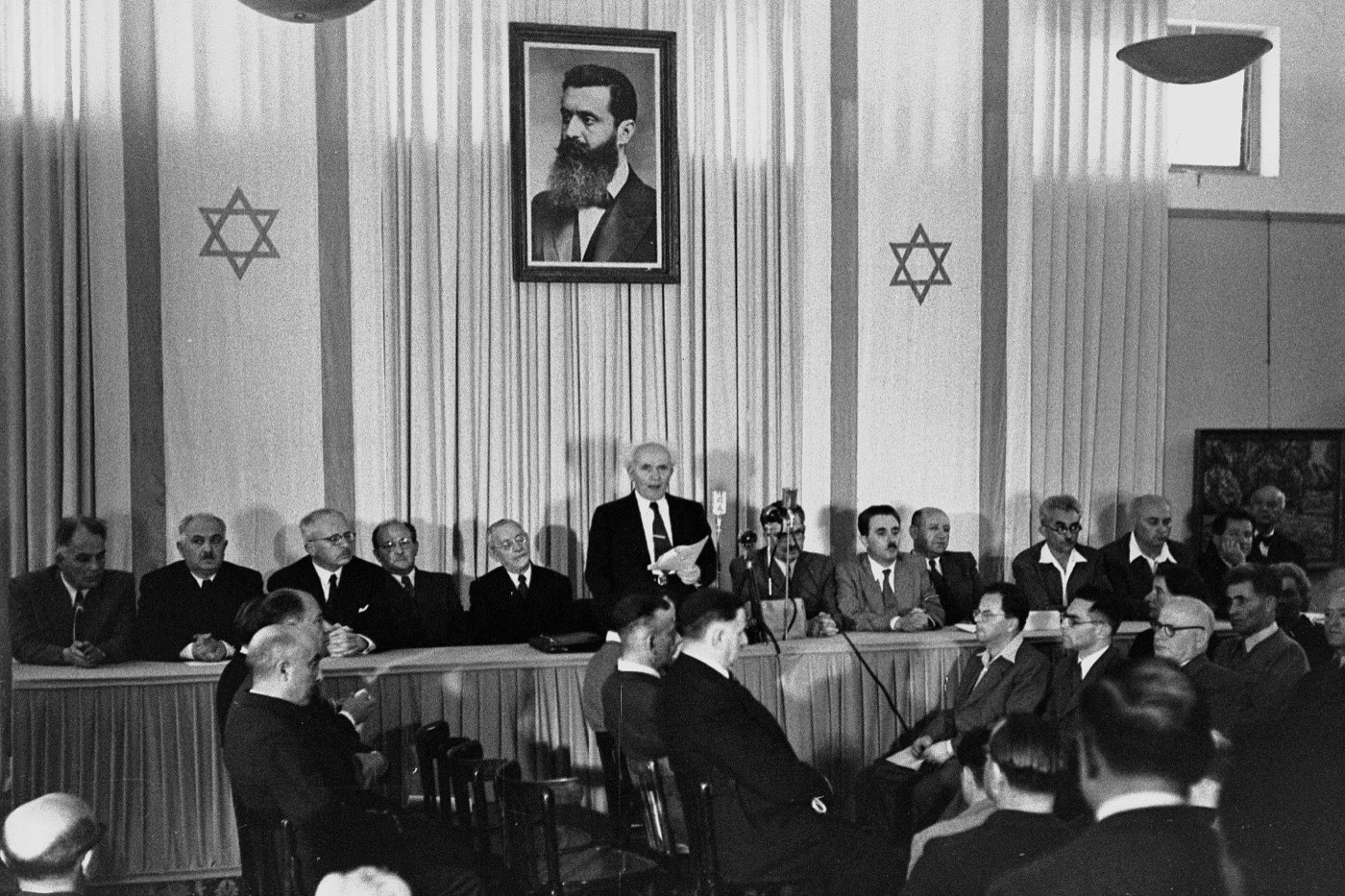 David Ben-Gurion reading the Declaration of Independence of the State of Israel on 14 May 1948 in Tel Aviv, beneath a portrait of Theodor Herzl